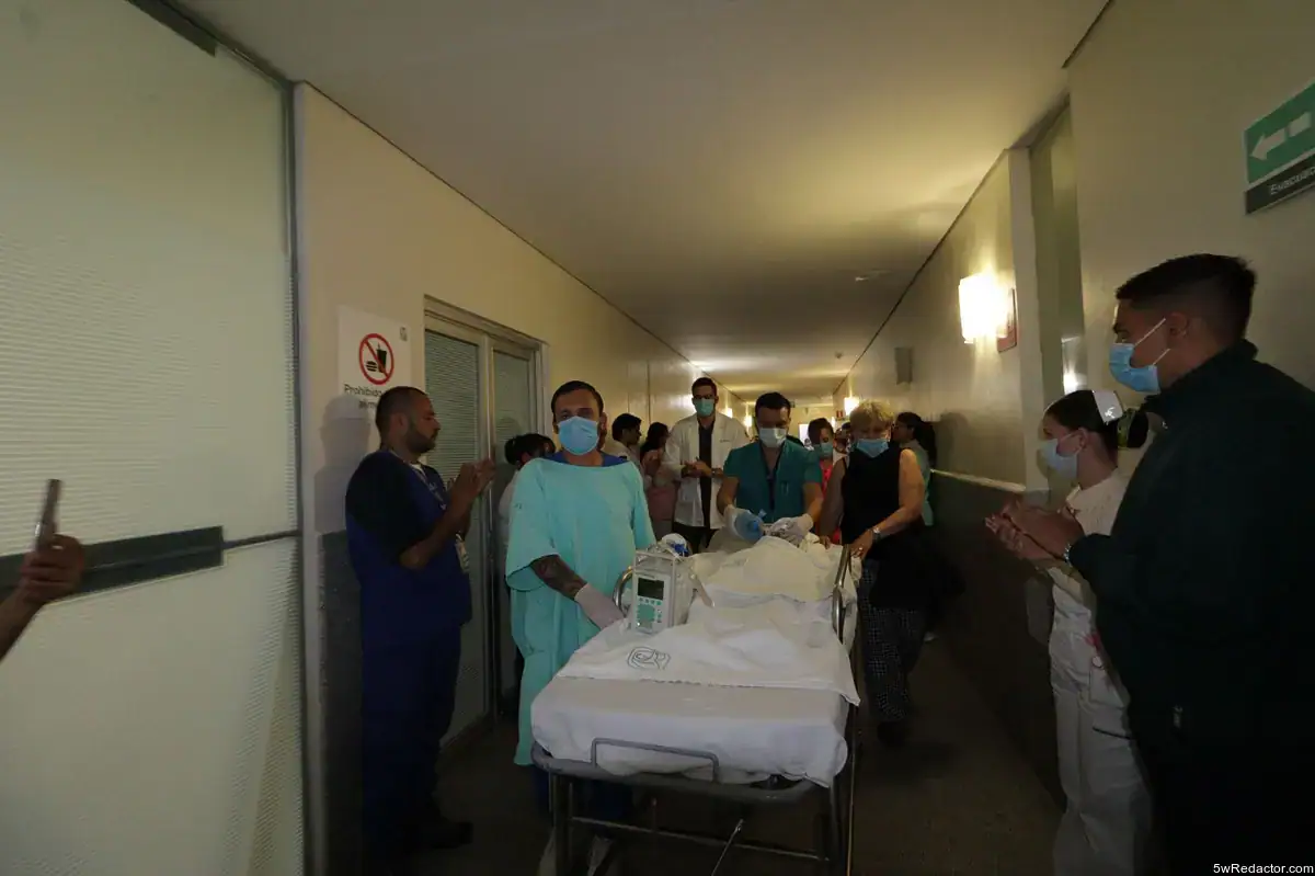 Personas en hospital con equipo médico y camilla, atención médica en pasillo, uso de mascarillas.